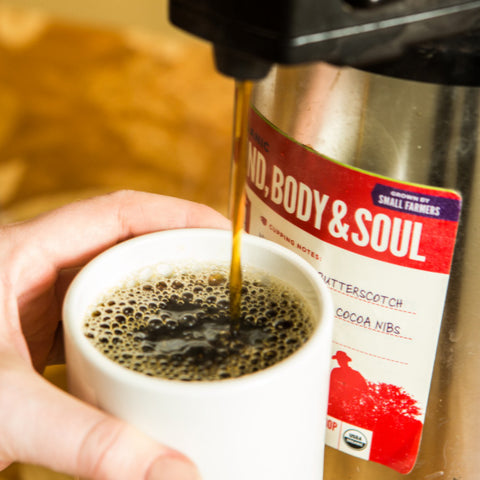 Filling a cup of coffee from an airpot labeled with Mind, Body & Soul coffee